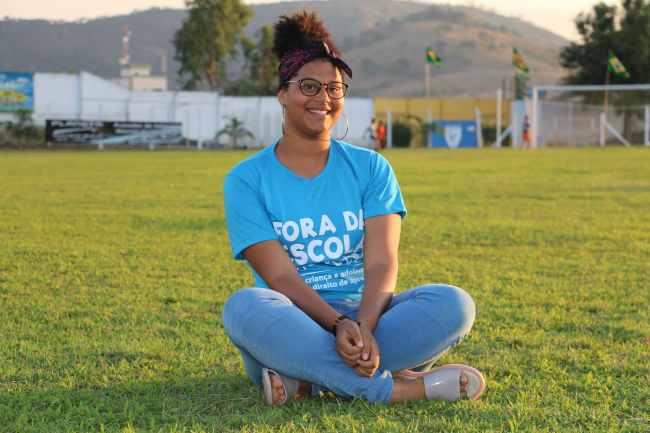 Adolescente sentada num gramado verde sorri para cÃ¢mera. Ela veste calÃ§a jeans azul e uma blusa azul. Ãculos de grau redondos, brincos de argola e uma faixa roxa e preta no cabelo 