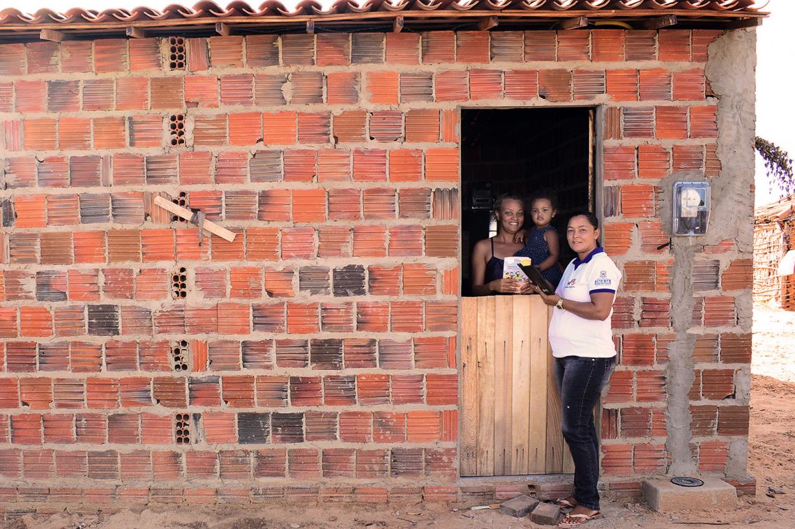 Uma agente, de blusa branca, estÃ¡ na porta de uma casa de tijolos. Ao seu lado estÃ¡ uma mulher negra jovem segurando seu filho, bebÃª, e sorrindo para a cÃ¢mera