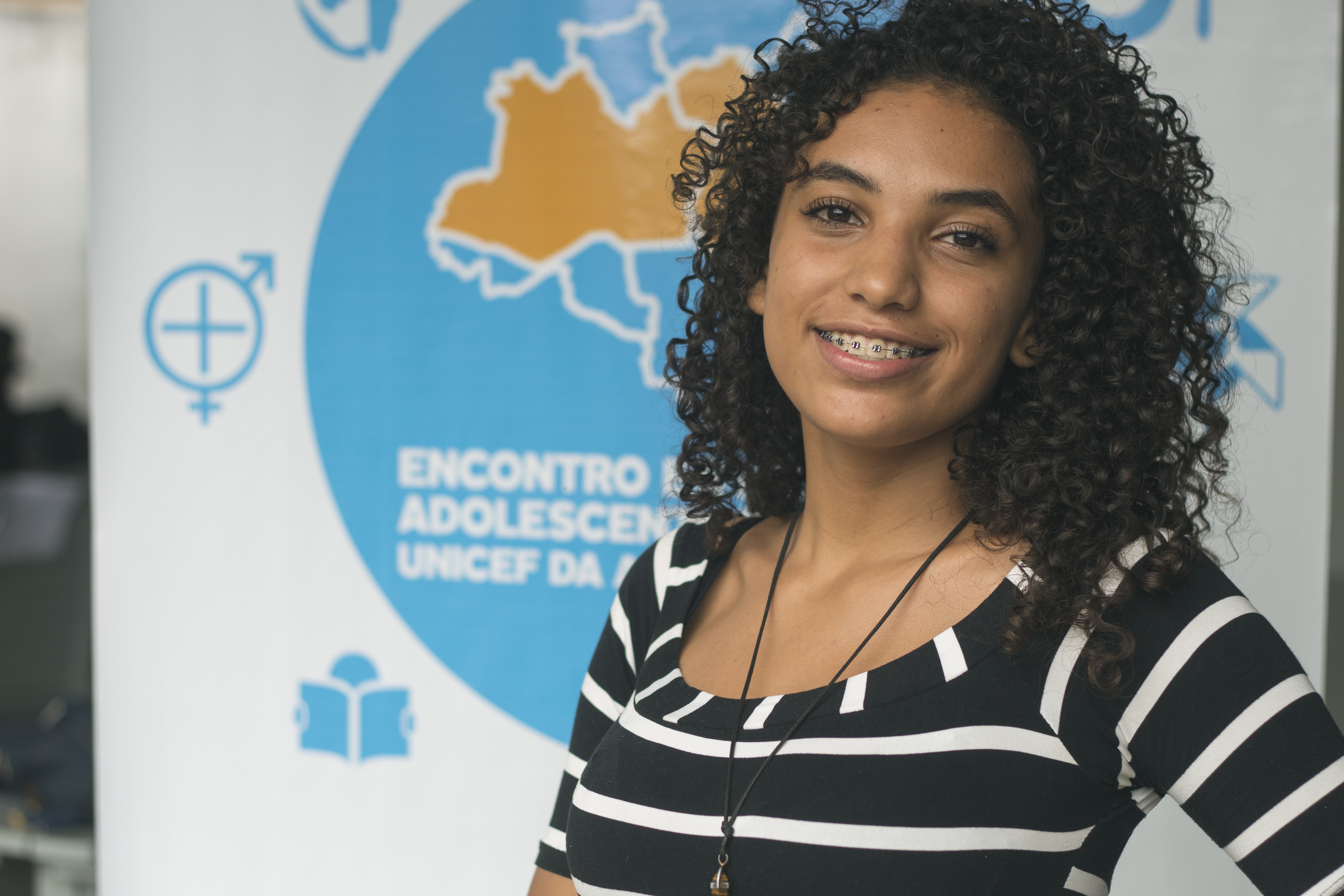 Jovem olha para frente e sorri. Ela veste blusa preta com listras brancas, cabelo solto e usa aparelho nos dentes. Ao fundo, um painel branco com detalhes em azul indica que ali é um encontro de adolescentes do NUCA