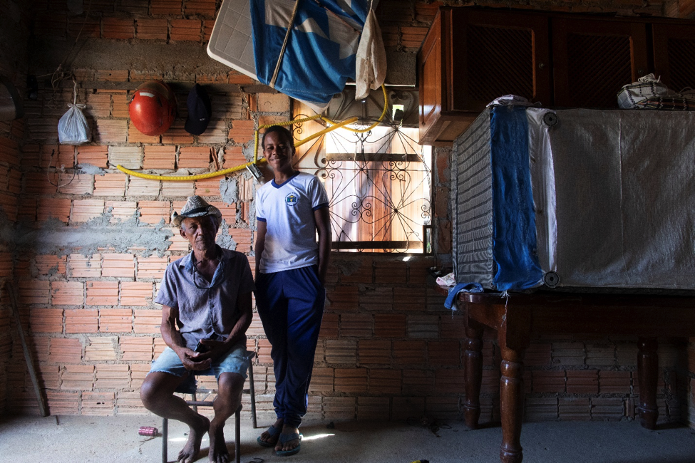 Adolescente vestindo uniforme escolar estÃ¡ em pÃ© sorrindo ao lado de um senhor de idade sentado numa cadeira, que veste bermuda jeans, camisa azul desbotada e chapÃ©u branco com fita preta. Eles estÃ£o na sala de uma casa humilde, com parede de tijolos Ã  mostra e chÃ£o sem revestimento. Do lado direito, encima de uma mesa de madeira estÃ¡ um objeto que parece ser uma cama virada. Na parede atrÃ¡s deles, estÃ£o pendurados um capacete vermelho, um bonÃ© preto e um saco plÃ¡stico