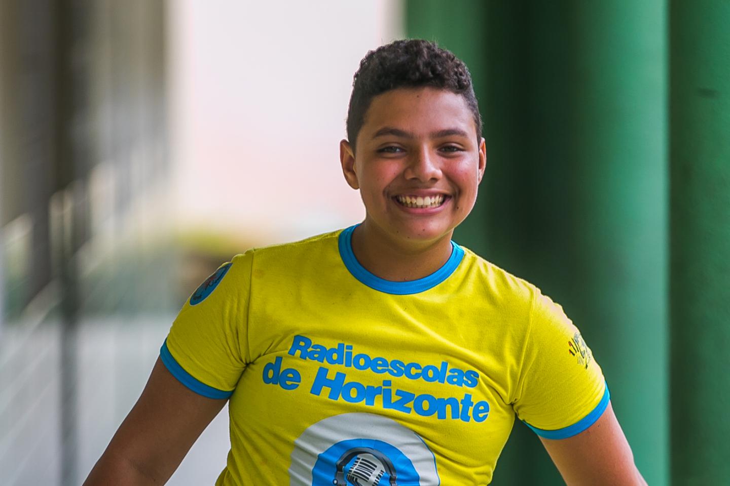 Adolescente sorrindo olha para a cÃ¢mera. Veste camiseta amarela fosforescente com detalhes em azul na manga. A estampa diz: "Radioescolas de Horizonte"  