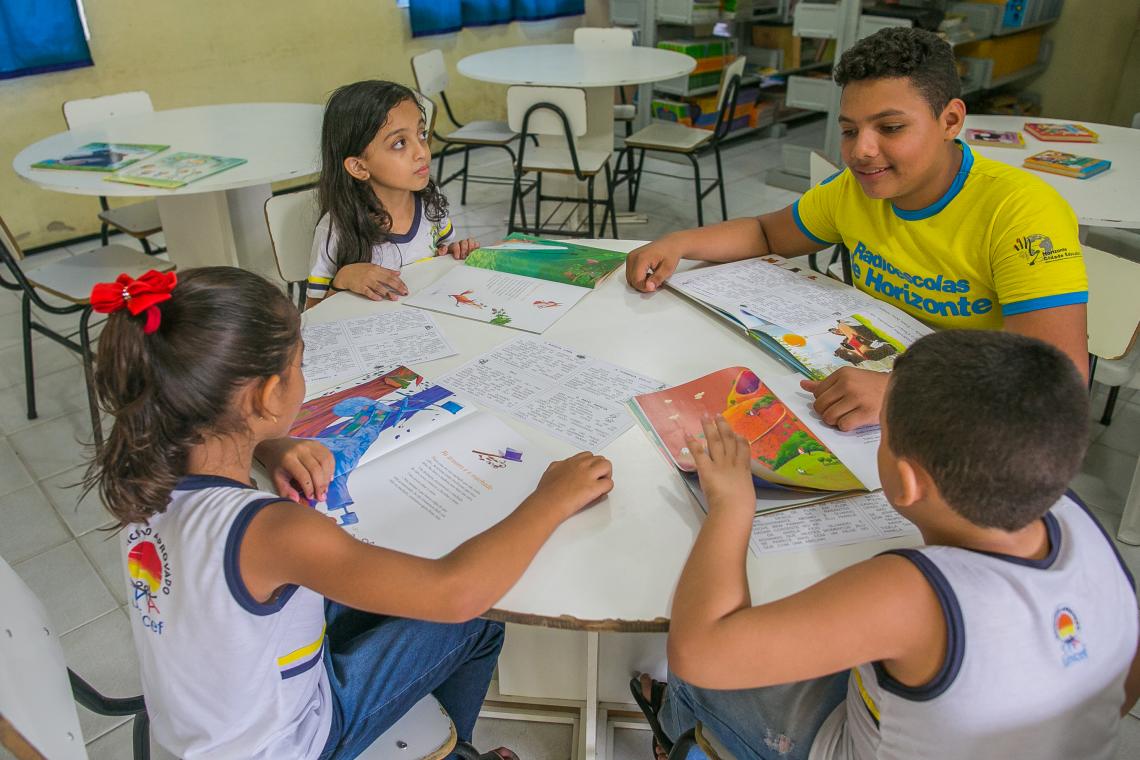 AcÃ¡ssio vestindo uma camiseta amarela fosforescente estÃ¡ sentado numa mesa redonda. Ao redor da mesa, trÃªs crianÃ§as menores de 12 anos vestem uniforme escolar e seguram livros nas mÃ£os. A mesa estÃ¡ repleta de livros. Ao fundo, estantes com livros indicam que estÃ£o numa biblioteca escolar