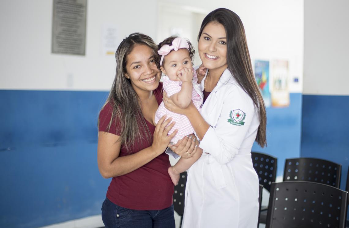 A imagem mostra Maria de FÃ¡tima (mÃ£e) e uma mÃ©dica, que utiliza jaleco branco, segurando JÃºlia (filha), que estÃ¡ entre as duas mulheres. As trÃªs estÃ£o em uma recepÃ§Ã£o de um ambiente hospitalar.