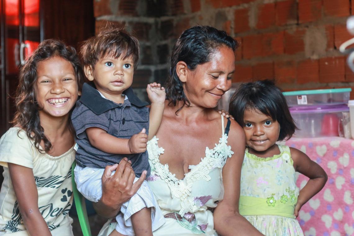 Mulher segurando um bebÃª de um ano, sorri para a filha de quatro anos, em pÃ© do seu lado direito. Do outro lado, a filha mais velha sorri para a cÃ¢mera