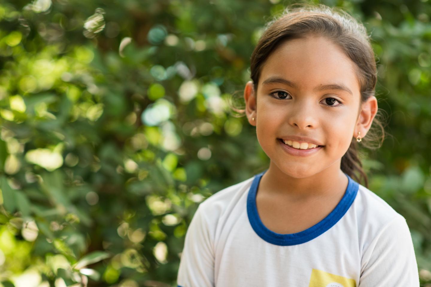 CrianÃ§a com uniforme escolar branco de gola azul olha para a cÃ¢mera e sorri. Ao fundo, temos uma vegetaÃ§Ã£o verde