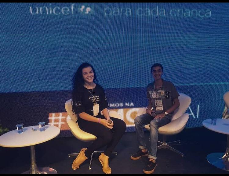 Adolescente vestindo blusa e calÃ§a preta sorri. Ela segura um microfone nas mÃ£os e estÃ¡ sentada numa cadeira em cima de um palco. ao fundo, um painel do UNICEF. Ao lado dela, outro jovem sentado numa cadeira sorri