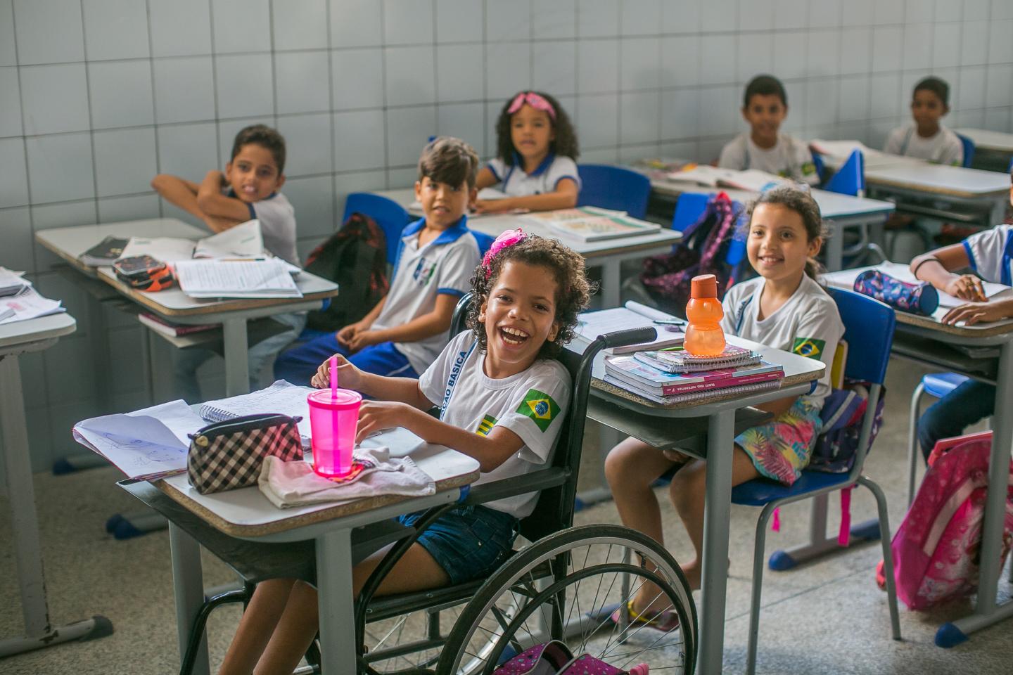 Vestindo uniforme escolar, menina sorri para cÃ¢mera. Ela estÃ¡ numa cadeira de rodas encaixada na carteira escolar. Sobre a mesa, um estojo quadriculado preto e branco, uma garrafinha de Ã¡gua rosa e um caderno. Ao fundo da menina, outras crianÃ§as estÃ£o sentadas em suas carteiras vestindo uniforme escolar e olhando para frente