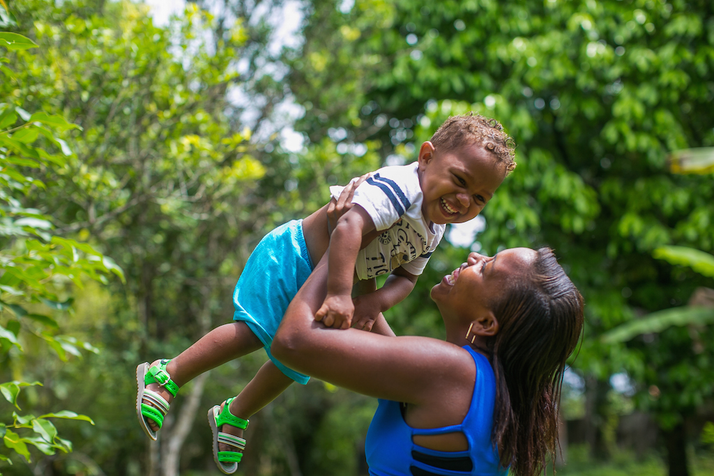 Mulher quilombola veste blusa azul e sorri para o filho pequeno. Ela o ergue. O menininho sorri, suspenso no ar. Ele veste uma blusa branca com listras azuis escuras e um desenho amarelo no peito, bermuda azul claro e sandalhas verdes fosforescentes. Ao fundo, uma vegetaÃ§Ã£o verde