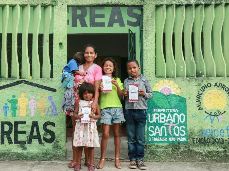 Mulher com vestido rosa segura um bebê no colo. O bebê está virado de costas para a câmera. Na frente e ao lado da mãe, seus outros 3 filhos olham para a câmera. As crianças seguram nas mãos um panfleto do governo municipal. Eles estão na porta de um CREAS, o Centro de Referência em Assistência Social. No muro verde do edifício, estão pintados o logo do CREAS, do município de Urbano Santos e do Selo UNICEF