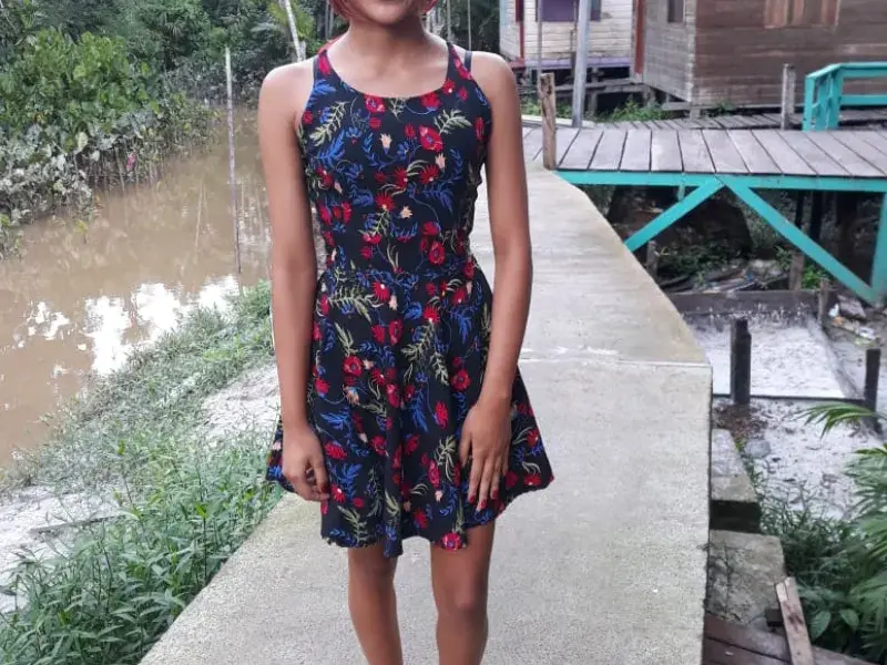 Adolescente com vestido preto com estampas florais sorri para a câmera. Ela tem cabelos tingidos de vermelho e veste um diadema de flores vermelhas e brancas. Ao fundo, casas de palafita