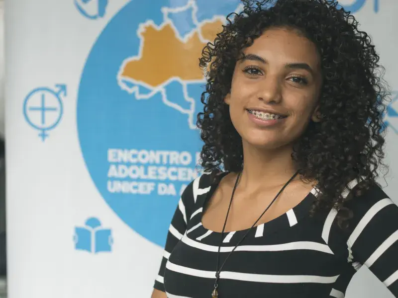 Jovem olha para frente e sorri. Ela veste blusa preta com listras brancas, cabelo solto e usa aparelho nos dentes. Ao fundo, um painel branco com detalhes em azul indica que ali é um encontro de adolescentes do NUCA