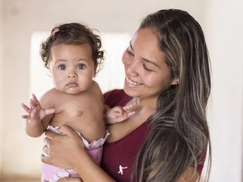 Maria de Fátima, uma mulher jovem utilizando uma blusa vermelha, segura e olha para sua filha Júlia. Júlia, que é uma bebê, está com um dos braços levantados, usa fralda rosa e olha para a câmera.