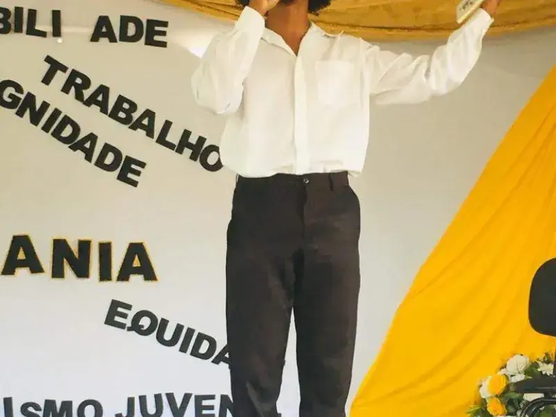 Jovem vestindo camisa branca e calça preta fala ao microfone em cima de um palco. Ele está com os braços abertos e segura em uma das mãos um livro