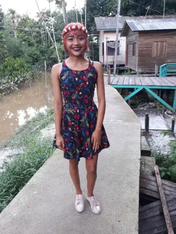 Adolescente com vestido preto com estampas florais sorri para a câmera. Ela tem cabelos tingidos de vermelho e veste um diadema de flores vermelhas e brancas. Ao fundo, casas de palafita